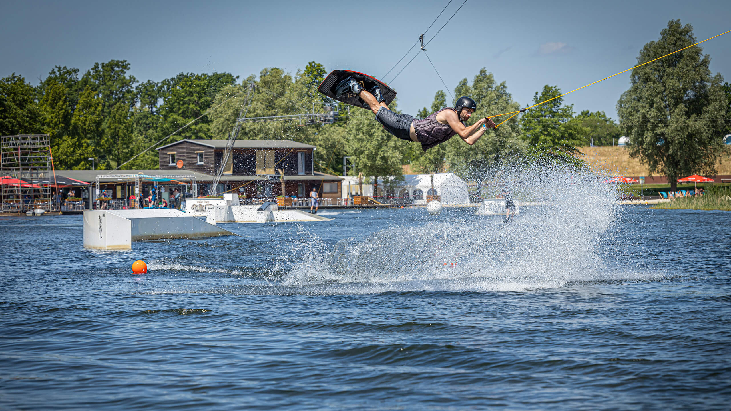  Wakeboardfahrer auf der Wakeboardanlage des Wakeparks Wolfsburg, fliegt über ein Hindernis, im Hintergrund die Gebäude des Parks.