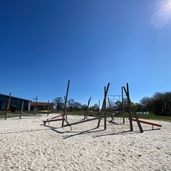 Spielplatz auf Sand mit Holzspielgeräten zum Klettern und Balancieren, im Hintergrund Gebäude des Badeland.