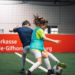 Ein Junge und ein Mädchen beim Zweikampf mit einem Fußball auf einem Indoor Fußballplatz des VfL Bolzwerk.