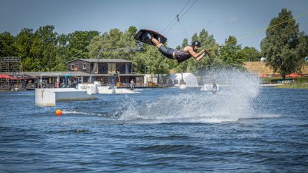  Wakeboardfahrer auf der Wakeboardanlage des Wakeparks Wolfsburg, fliegt über ein Hindernis, im Hintergrund die Gebäude des Parks.