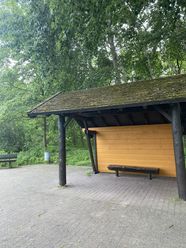 Überdachter Grillplatz mit einer Bank und Wald im Hintergrund.