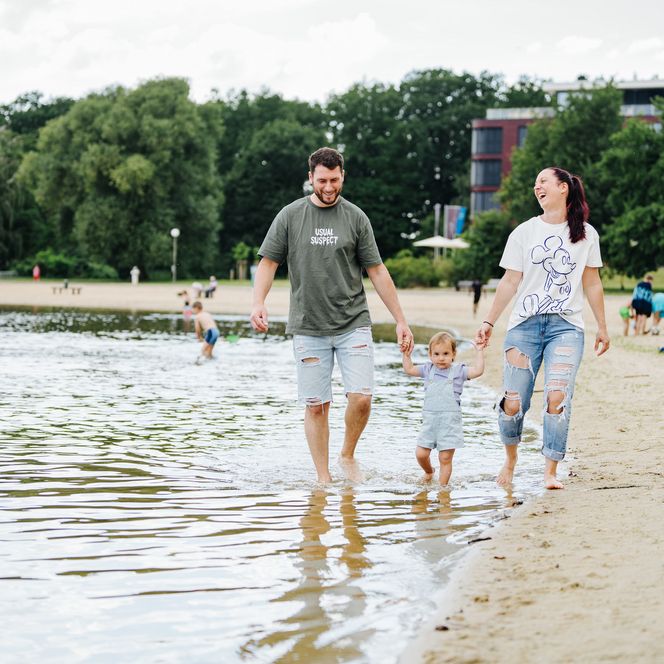 Eltern mit Kleinkind gehen am Strand des Allersees, mit den Füßen im Wasser, spazieren.